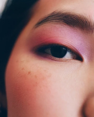 Close-up studio portrait of a woman's face features with violet and pink eyeshadow, defined eyebrow, and visible freckles on her cheek.