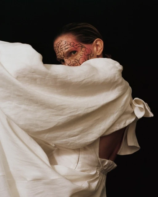 Woman with artistic face paint posing wrapped in a white sheet, against a dark background in a studio setting, eye-level shot.