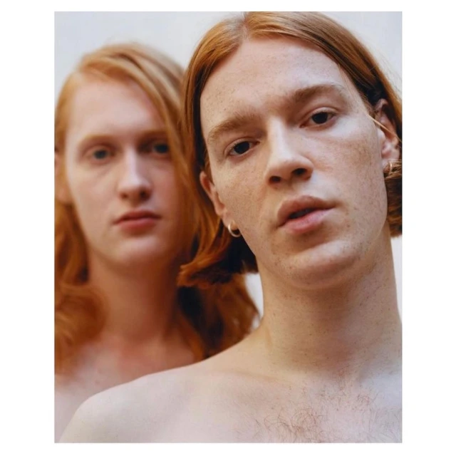 Close-up portrait of two fair-skinned, fair-haired, young adult males with reddish-blonde hair posing in a neutral indoor setting.