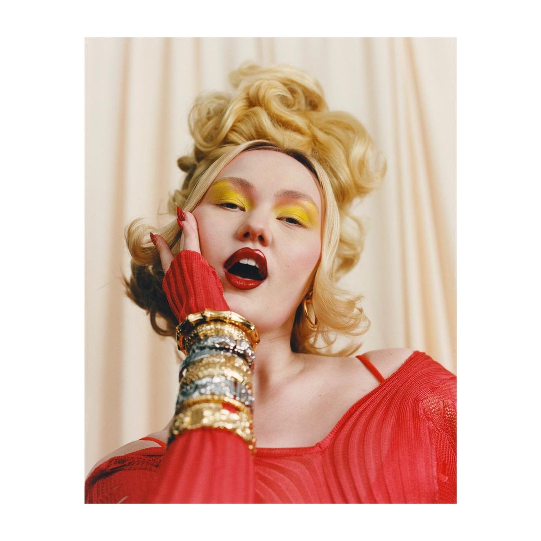 Headshot of a blonde adult female with bold yellow eye makeup and red lipstick, wearing a red top and silver and gold bracelets.