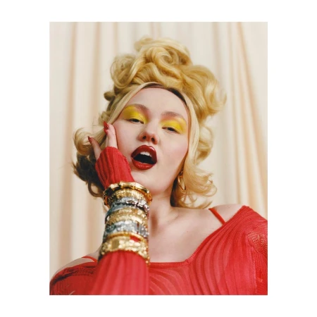 Headshot of a blonde adult female with bold yellow eye makeup and red lipstick, wearing a red top and silver and gold bracelets.