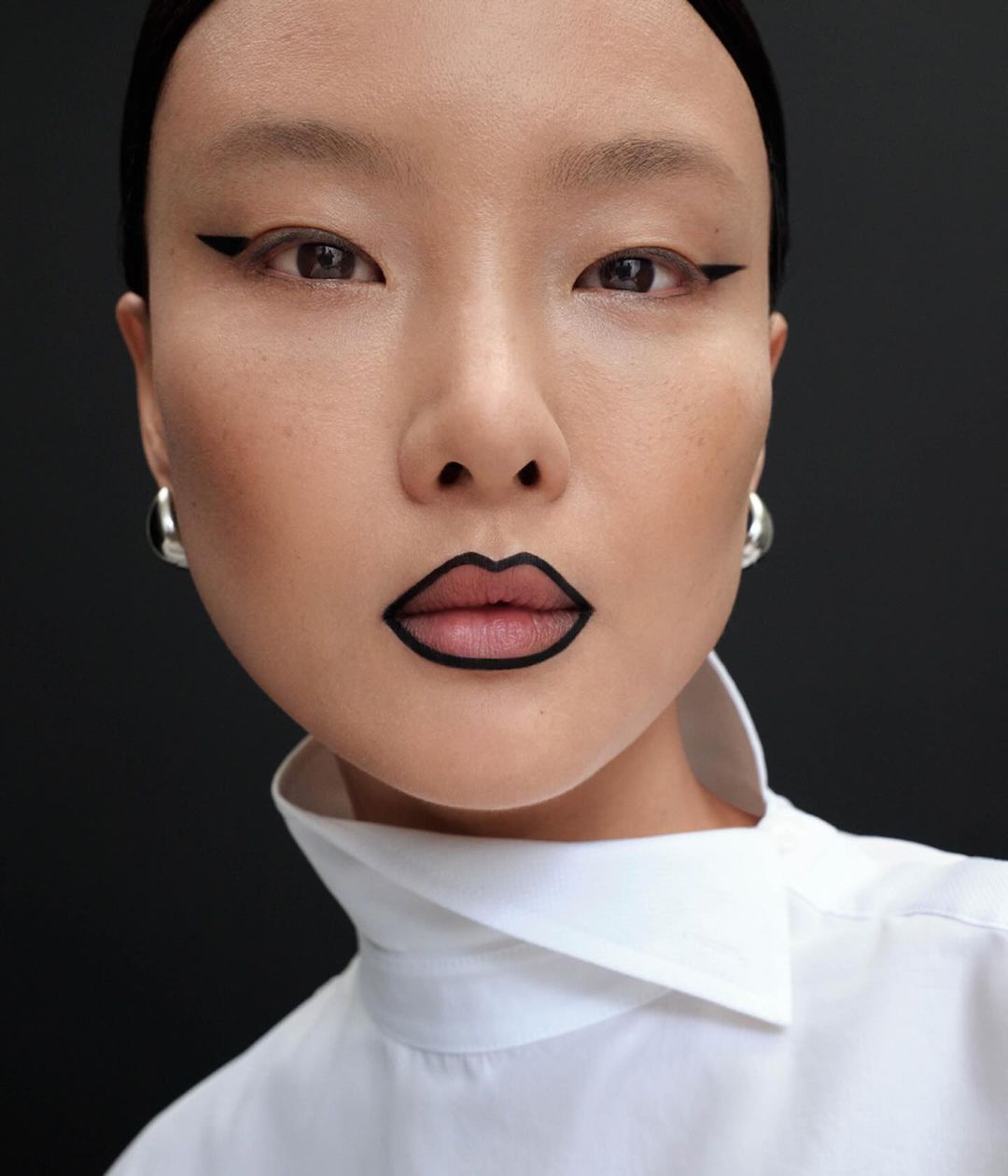 Close-up portrait of an East Asian model with graphic black lip liner and winged eyeliner wearing a white collared shirt.