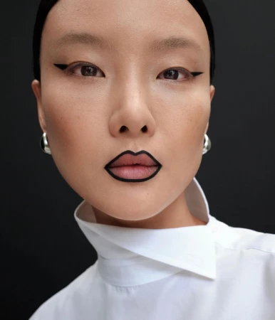 Close-up portrait of an East Asian model with graphic black lip liner and winged eyeliner wearing a white collared shirt.