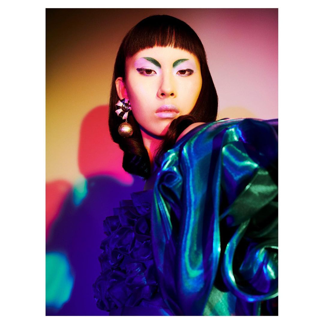 Portrait of Asian woman with dramatic makeup, wearing iridescent blue ruffled top, posing in front of a vibrant, colorful background.