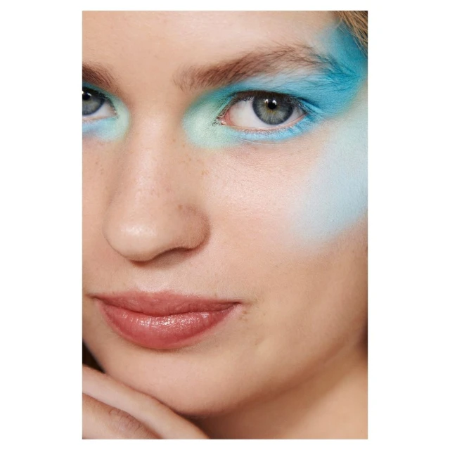 Close-up portrait of a Caucasian female model with blue and green eyeshadow posing in a brightly lit photo studio environment.