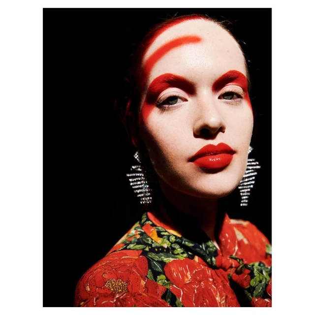 Close-up portrait of a woman with bold red makeup, including geometric patterns on her face, statement earrings, and a floral top.