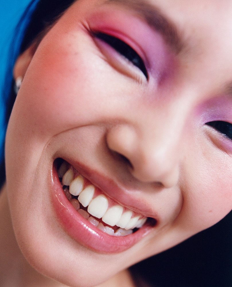 Close-up of a smiling Asian woman with purple and pink eyeshadow, showcasing beauty and makeup against a bright blue background.