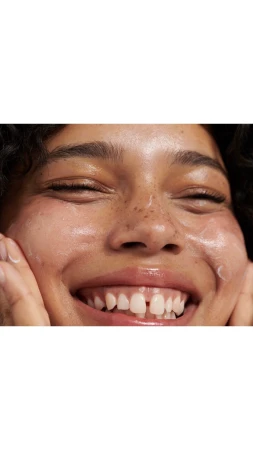 Close-up portrait of a smiling young person applying face cream with visible freckles and curly hair by Benjamin Madgwick