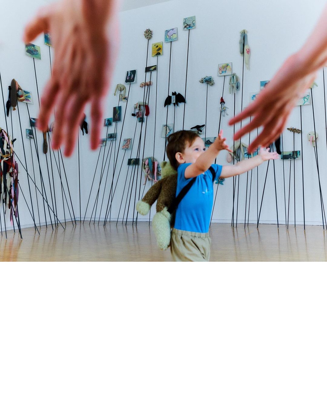 Toddler reaching for outstretched hands in an indoor setting with a teddy bear backpack and a display of whimsical artwork.