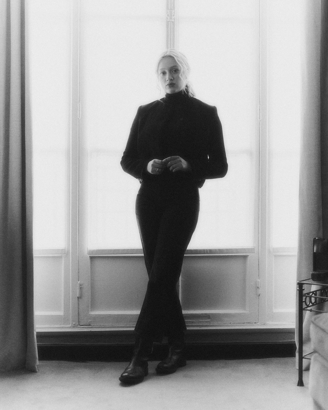 Elegant woman in black blazer and pants poses in front of a bright window in a neutral interior, creating a high-contrast black-and-white photo.
