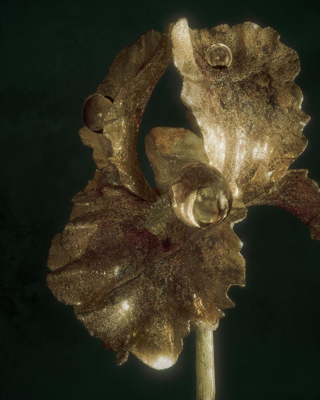 Close-up of a golden flower sculpture against a dark green background, showcasing intricate petal details and subtle metallic textures.