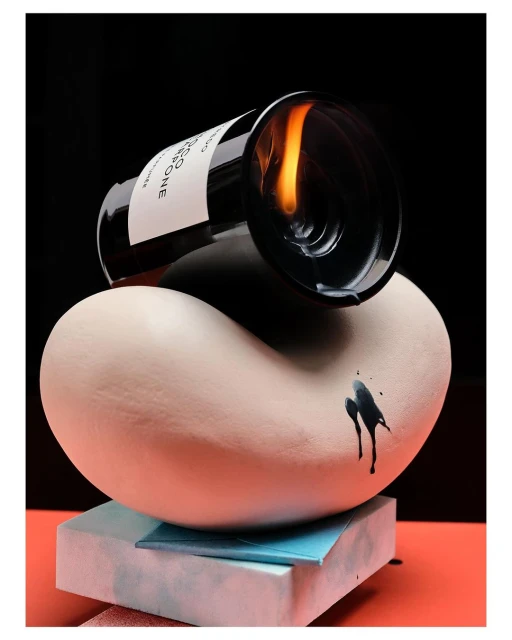 Conceptual still life featuring an open bottle on a tan sculpture, with black liquid drips and a fire reflection inside the bottle.