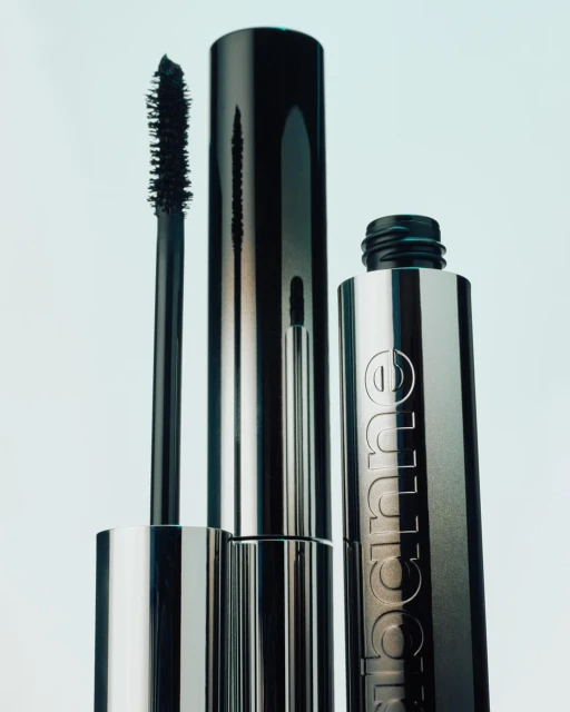 Close-up of silver mascara tubes and applicator with black mascara, against a bright backdrop in a studio setting.
