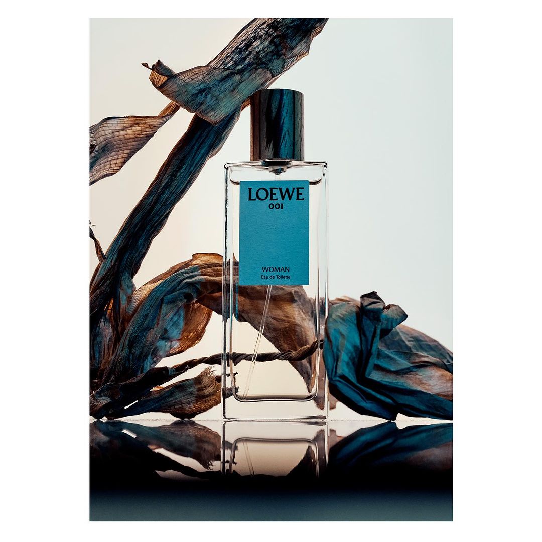 Loewe 001 Woman eau de toilette bottle stands out against dried teal botanicals, a reflective surface, and studio lighting.