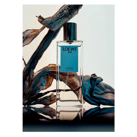 Loewe 001 Woman eau de toilette bottle stands out against dried teal botanicals, a reflective surface, and studio lighting.