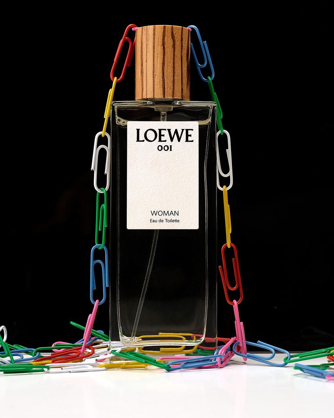Loewe 001 Woman eau de toilette fragrance bottle on a white surface, surrounded by colorful paper clips against a black background.