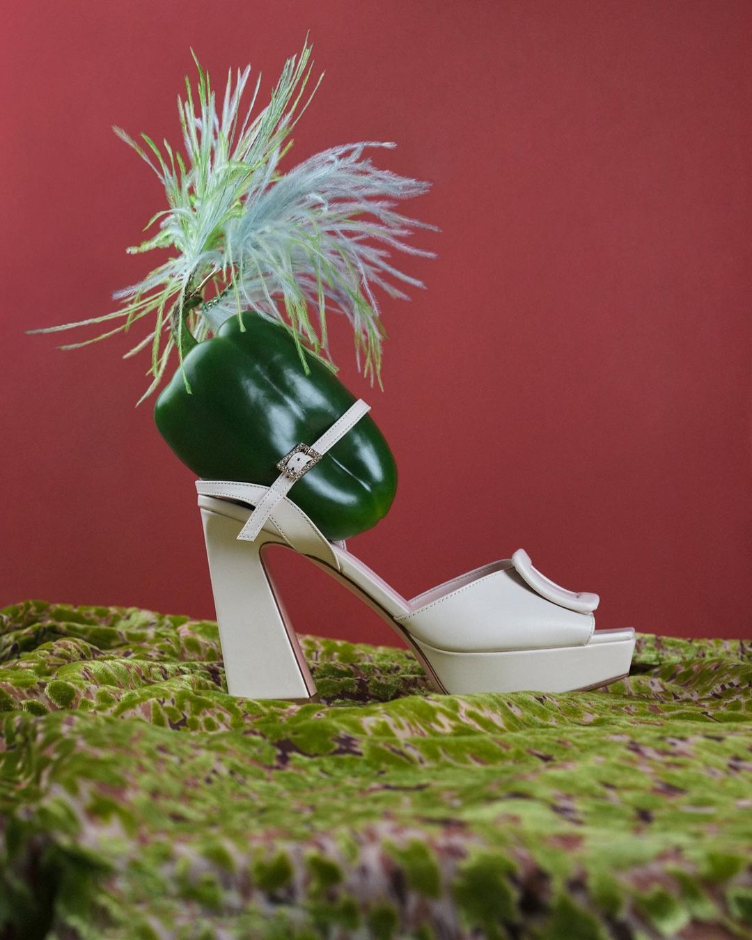 Fashionable sculptural sandal adorned with green bell pepper and feathered accents against red backdrop on decorative textile surface.