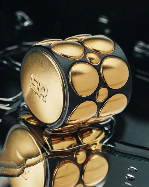 Close-up of a black and gold geometric object with rounded square and circular elements, resting on a reflective surface.