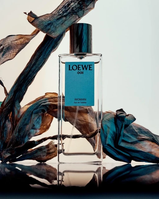 Loewe 001 Woman Eau de Toilette perfume bottle against a white background with dried floral elements and reflective surface.