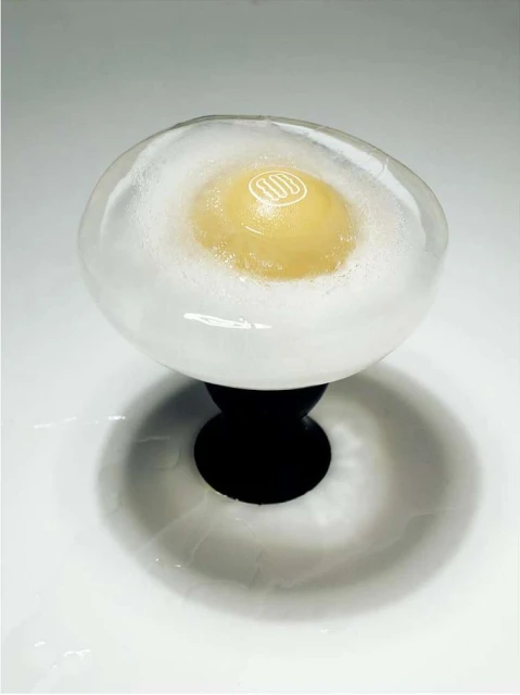 Close-up of a transparent egg-shaped orb on a black pedestal, filled with a yellow yolk-like center surrounded by white, foamy material.