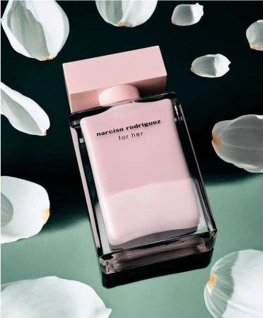 A rectangular pink perfume bottle with a matching cap surrounded by scattered white flower petals on a dark reflective surface