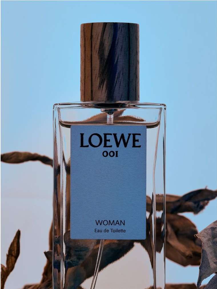 Close-up of a Loewe 001 Woman Eau de Toilette bottle, showcasing its transparent glass, wooden cap, and light blue product label.