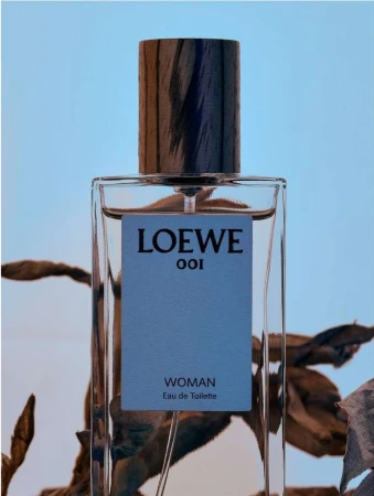 Close-up of a Loewe 001 Woman Eau de Toilette bottle, showcasing its transparent glass, wooden cap, and light blue product label.