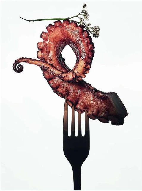 Close-up of grilled octopus tentacle artfully displayed on a fork, adorned with delicate white flowers against a clean white background.