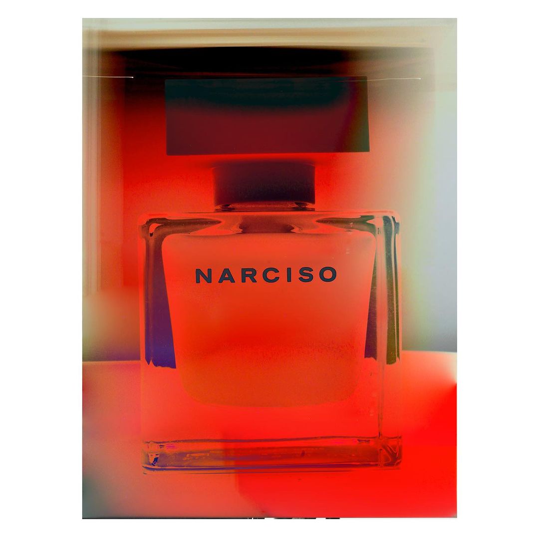Close-up of a transparent glass bottle of Narciso perfume, centered in the frame, with a dark cap and a bright orange glow.