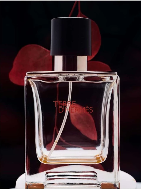 Close-up studio shot of a Terre D'Hermès fragrance bottle featuring clear glass and a black cap against a black background with red leaves.