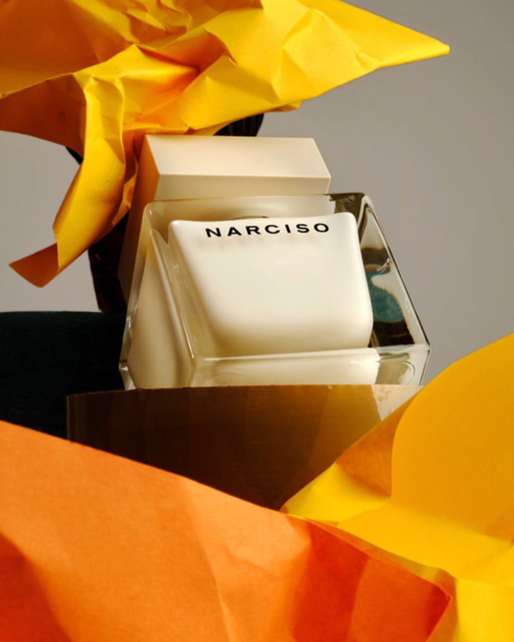 Close-up of a Narciso perfume bottle surrounded by colorful yellow and orange paper against a neutral background in studio light.