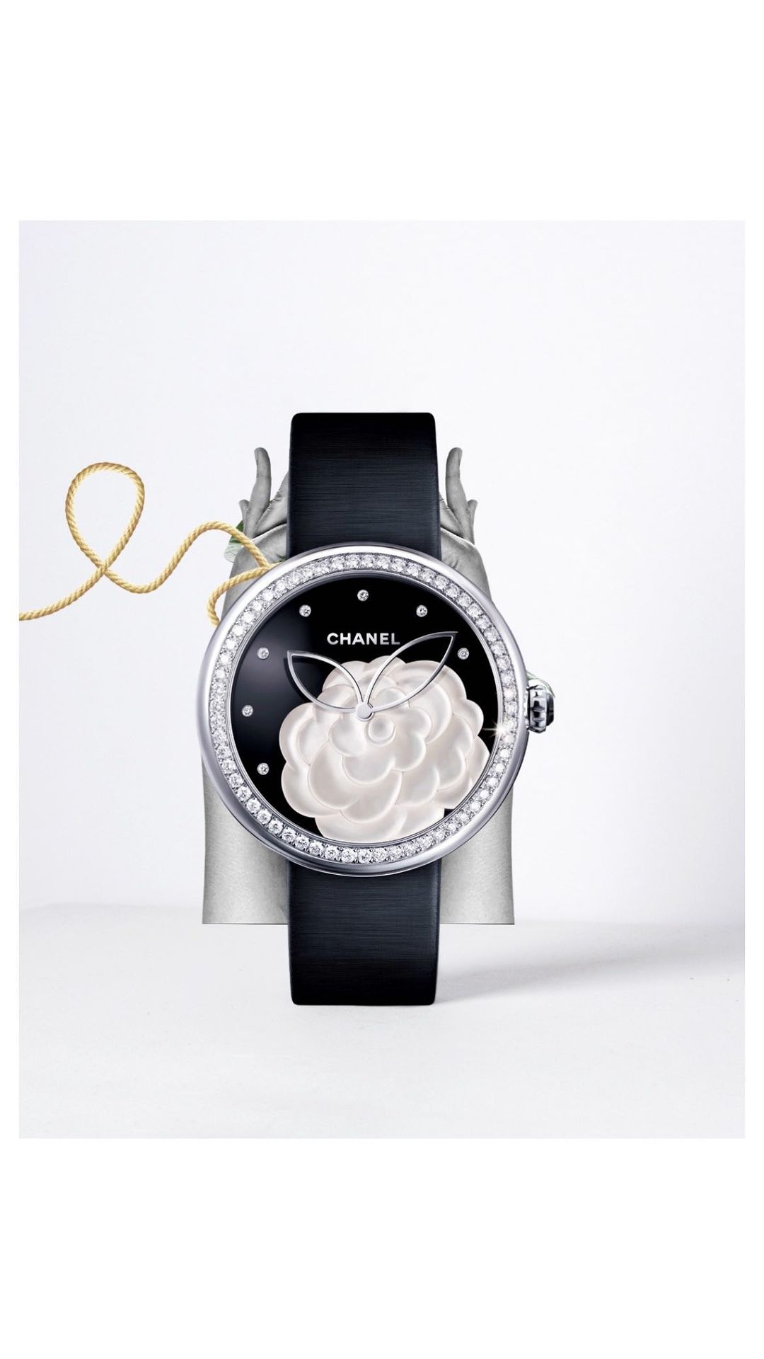 Elegant Chanel watch showcasing a black dial with a white camellia design, set against a light backdrop and silver stand, with a gold cord.