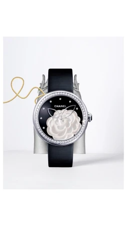 Elegant Chanel watch showcasing a black dial with a white camellia design, set against a light backdrop and silver stand, with a gold cord.