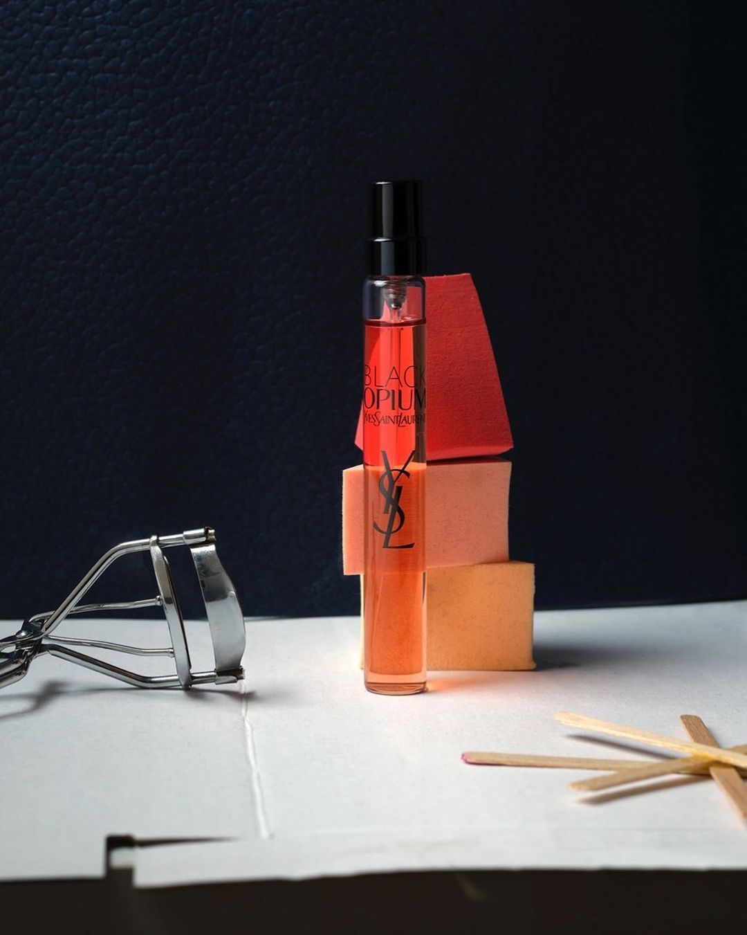 Still life shot of a miniature YSL Black Opium perfume bottle, beauty blender sponges, lash curler, and cosmetic applicator sticks.