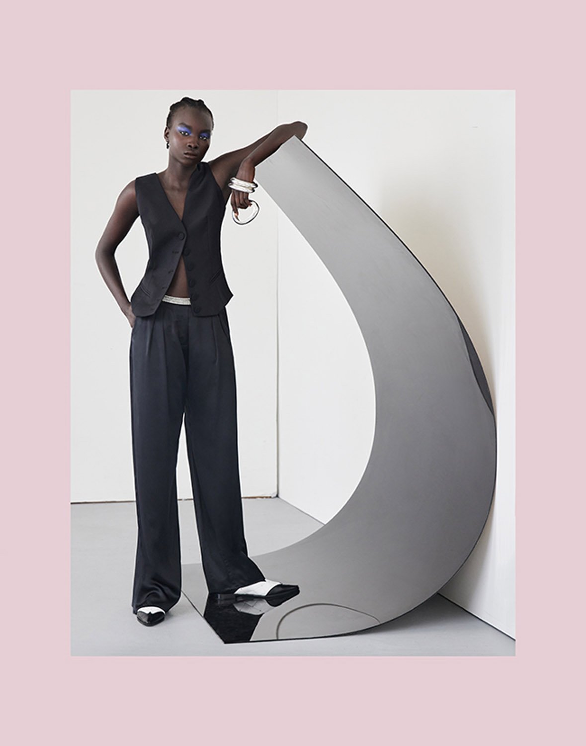 A Black model poses next to a gray sculpture in a studio setting. The model wears a black vest and wide-leg pants, with white-tipped shoes.