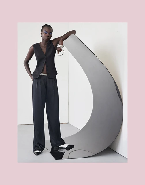 A Black model poses next to a gray sculpture in a studio setting. The model wears a black vest and wide-leg pants, with white-tipped shoes.