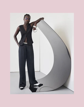 A Black model poses next to a gray sculpture in a studio setting. The model wears a black vest and wide-leg pants, with white-tipped shoes.