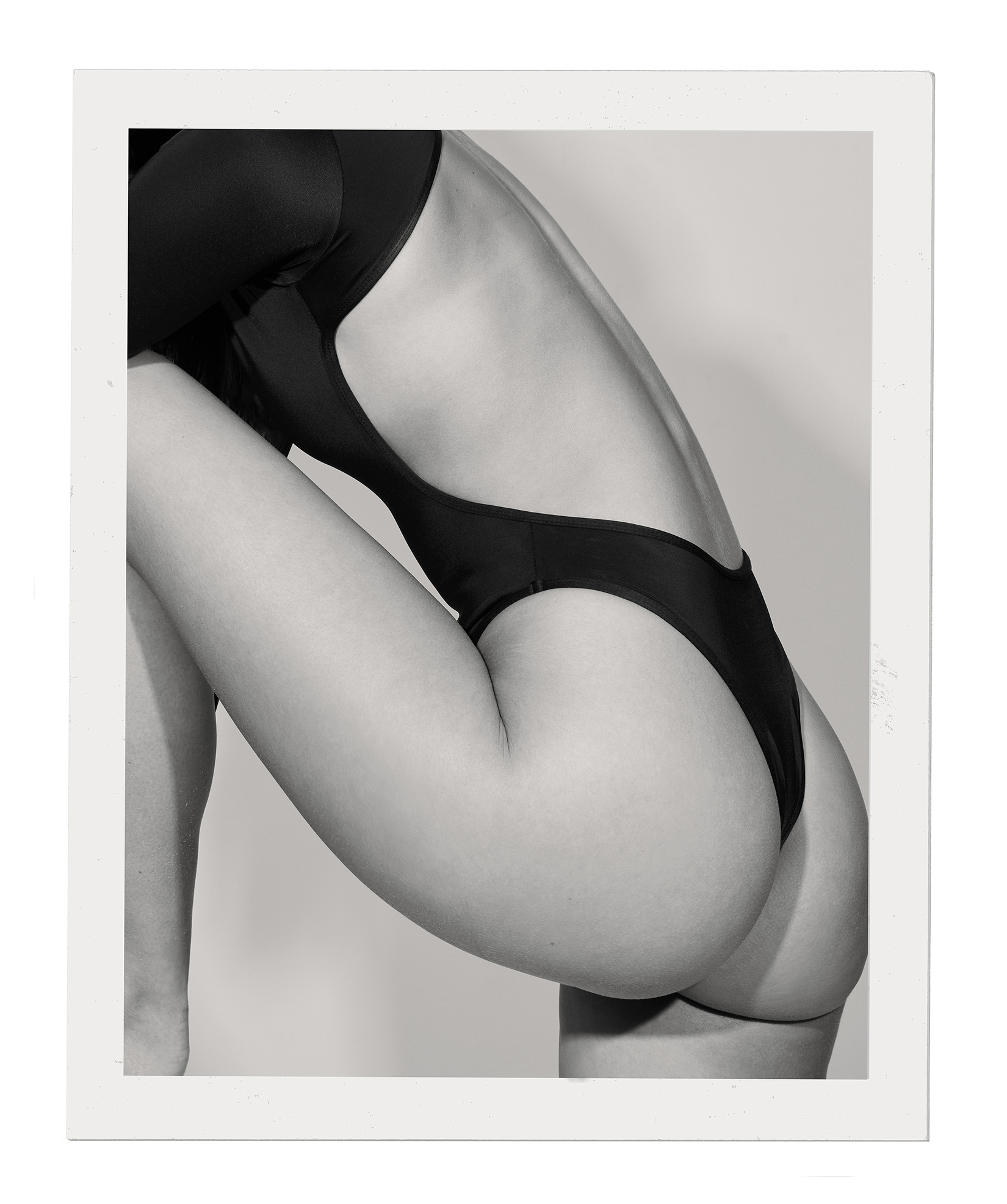 Studio shot of a nude woman in a black one-piece swimsuit with an arching back and legs, photographed in black and white with a plain backdrop.