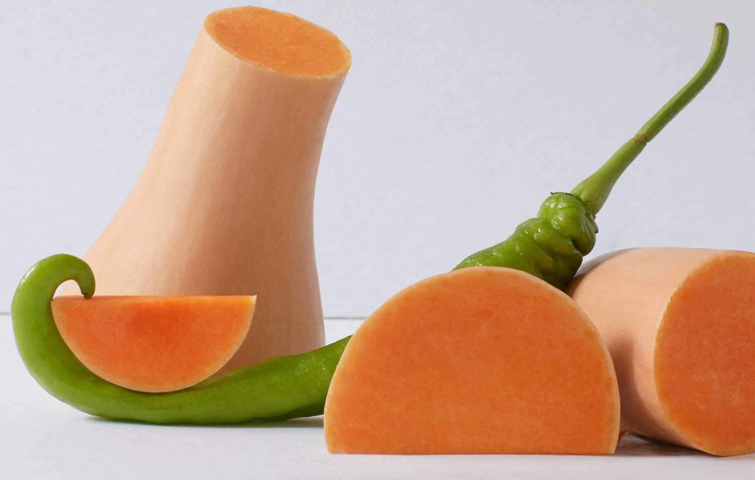 Still life studio shot of sliced butternut squash with a green chili pepper creating a curved line against a white background.