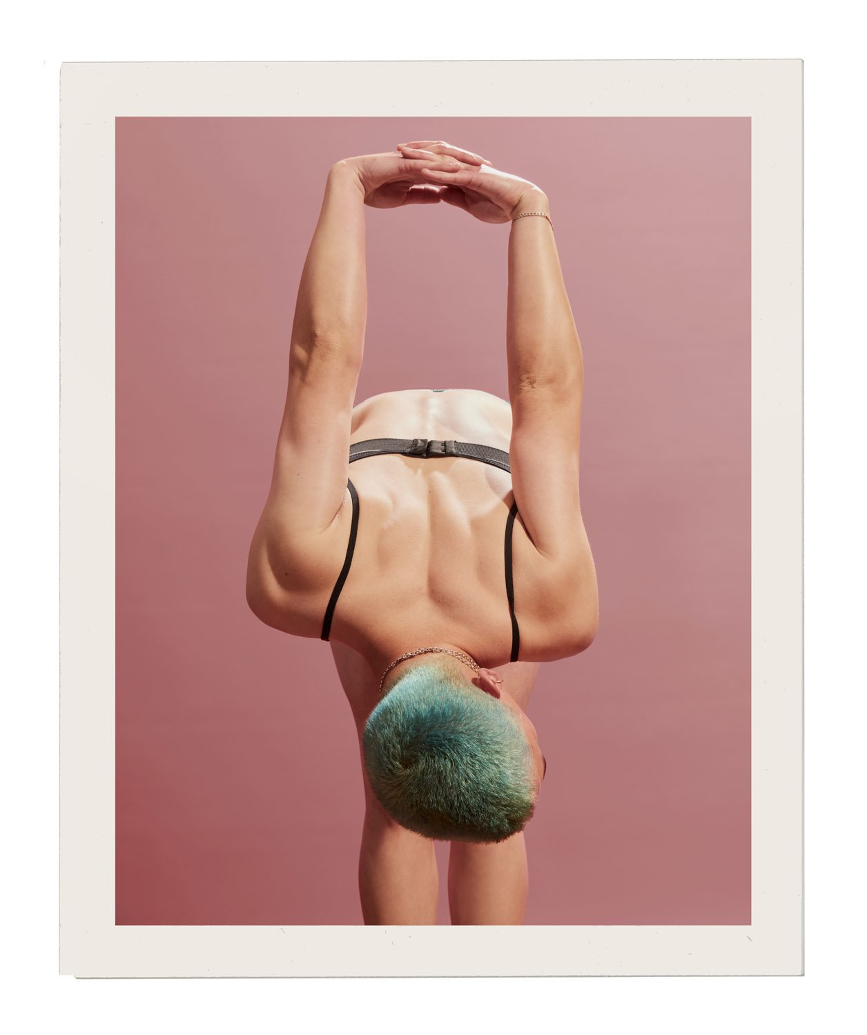 Back view of a slender adult female in a black sports bra, leaning forward with arms raised overhead against a dusty rose background.