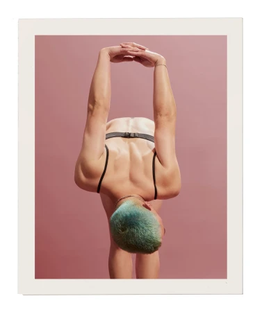 Back view of a slender adult female in a black sports bra, leaning forward with arms raised overhead against a dusty rose background.