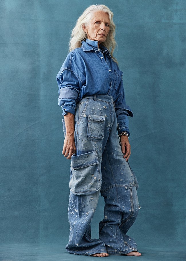 Older woman in denim button-down and distressed denim cargo pants poses in a studio setting with a textured blue background.