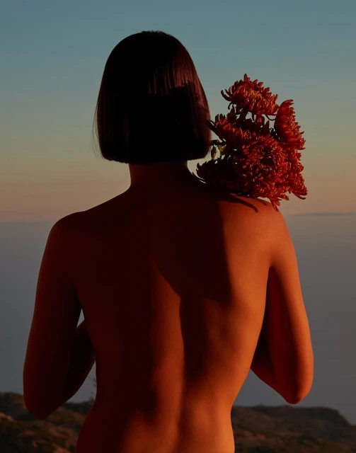 Back view of nude woman with short dark hair holding a bunch of orange chrysanthemums over her shoulder against a sky background.