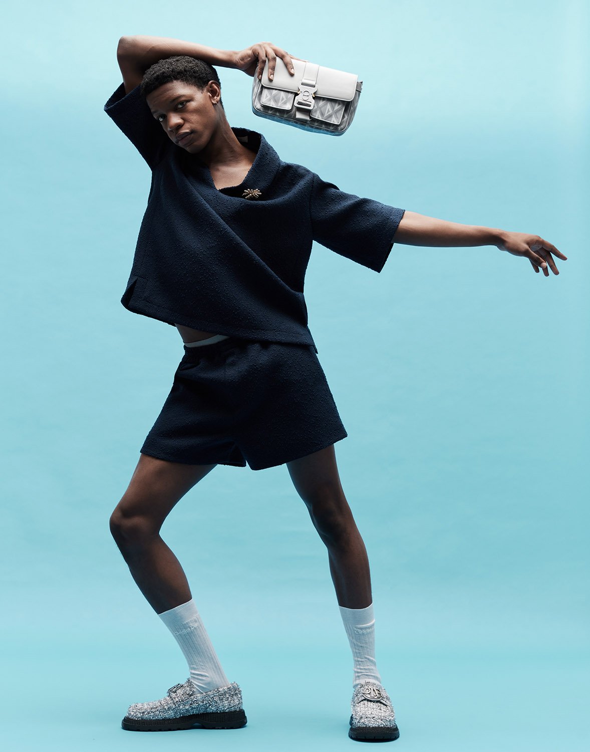 Fashion shot of a person wearing a navy blue outfit, white socks and patterned loafers, holding a purse against a blue backdrop.