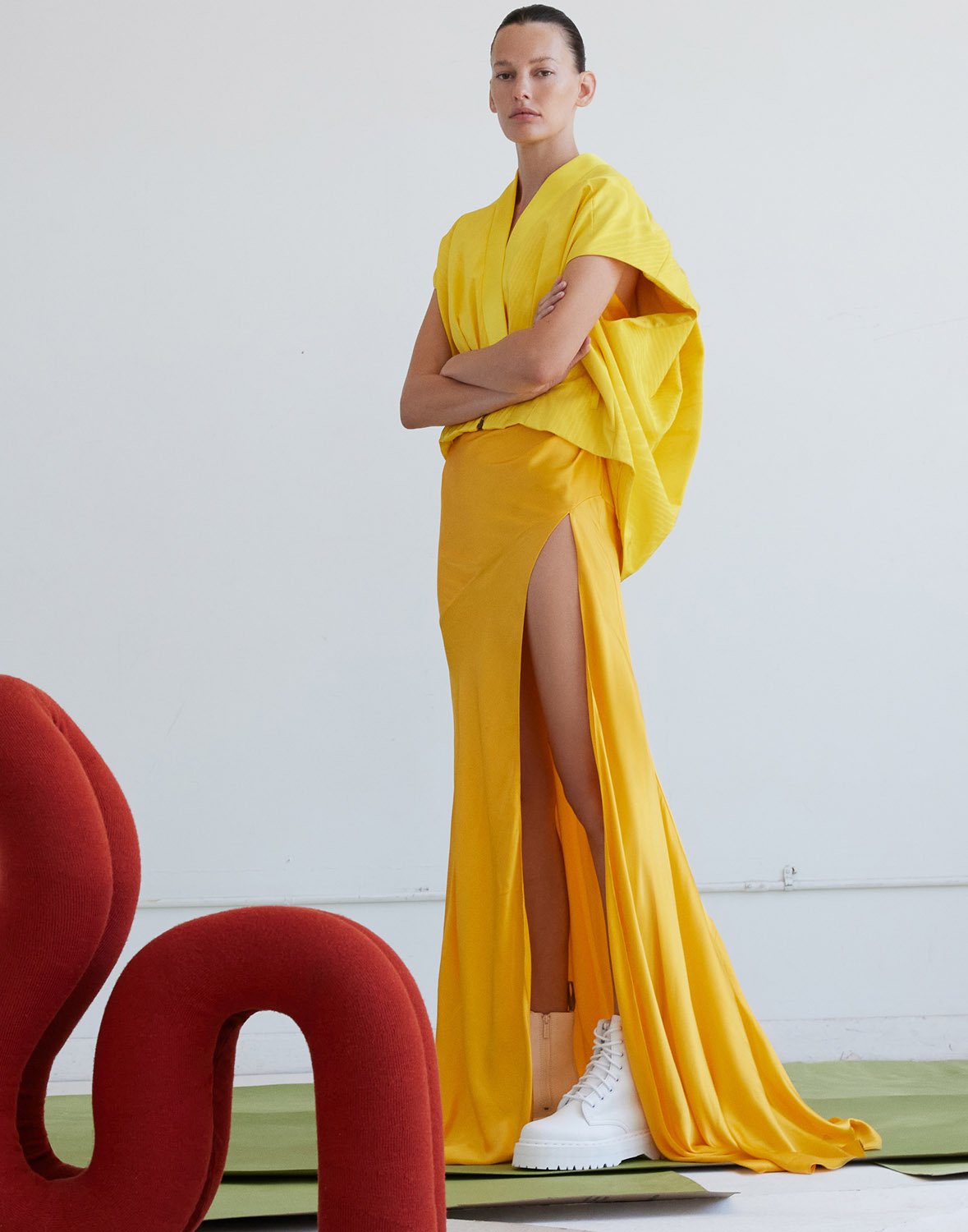 Woman in yellow gown with high slit poses in front of white background, wears white platform sneakers and red chair is visible.