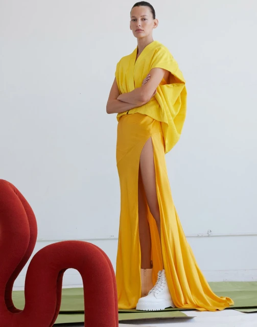 Woman in yellow gown with high slit poses in front of white background, wears white platform sneakers and red chair is visible.