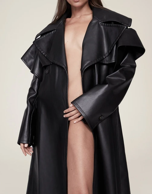 A fashion shot features a slender adult woman posing in a long, open black leather trench coat against an off-white background.