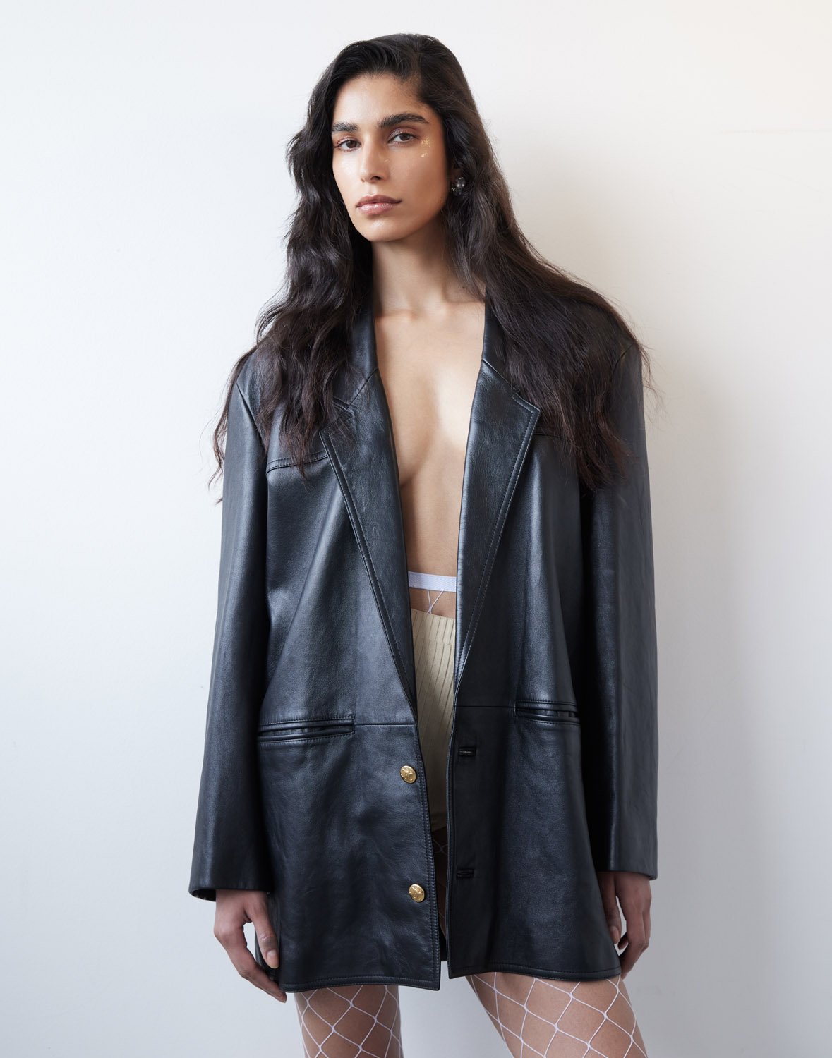 Model in an oversized black leather blazer wearing fishnet tights against a plain white background by Manolo Campion