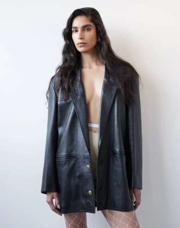 Model in an oversized black leather blazer wearing fishnet tights against a plain white background by Manolo Campion