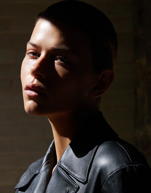 Close-up portrait of a young adult female model wearing a dark leather jacket, illuminated by directional natural light creating deep shadows.
